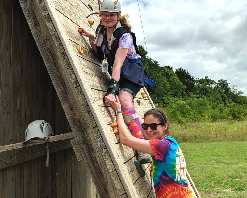 Kamp Kaleidoscope counselor helping camper climb rock wall